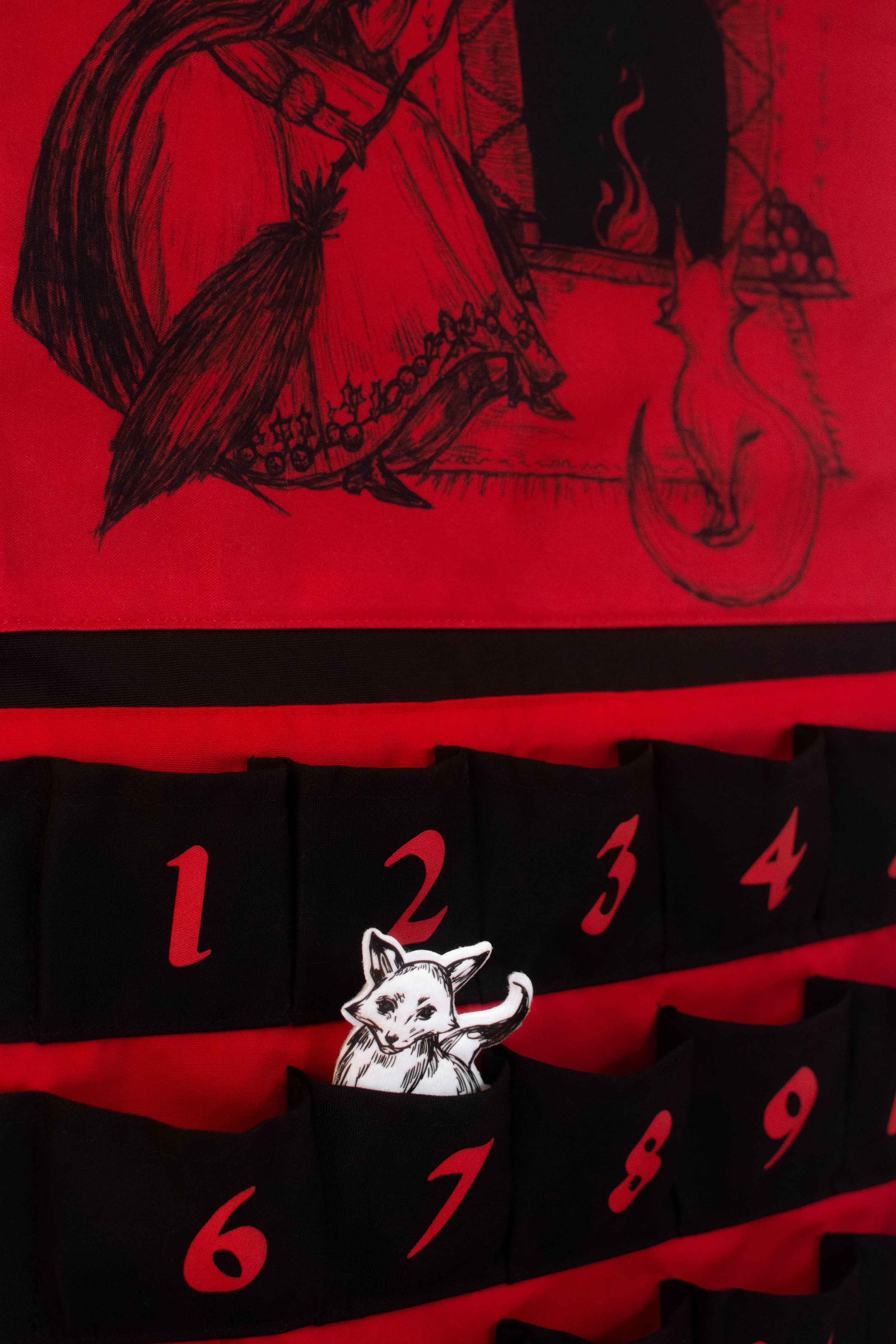Large black and red canvas advent calendar hanging from a wooden dowel and black rope. Featuring 25 pockets for the 25 days leading up to christmas. The top half is a large drawing of a witch and her familiar, a fox, sitting by a holiday themed fire. Foxblood logo in red at the bottom. A white fox market can be placed in the pockets to mark each day